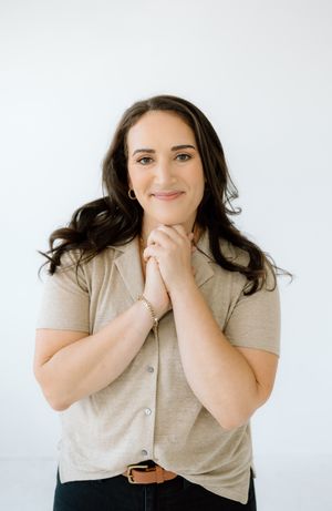 A professional headshot of someone in business casual attire smiling warmly with clasped hands.