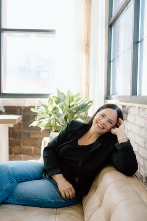 A casual portrait showing someone relaxing on a couch near a potted plant in natural window light.