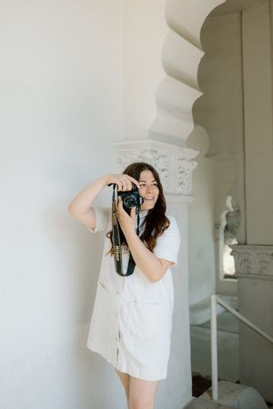 Woman in white dress taking photos with camera under architectural arches.