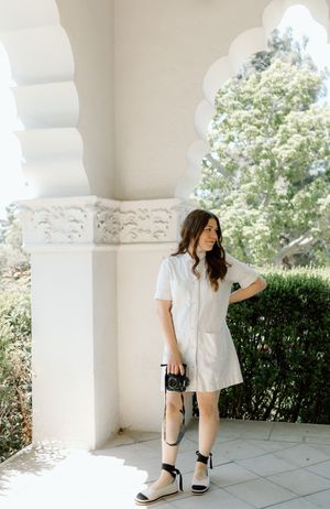 Woman in casual white dress posing with camera against ornate white archway.