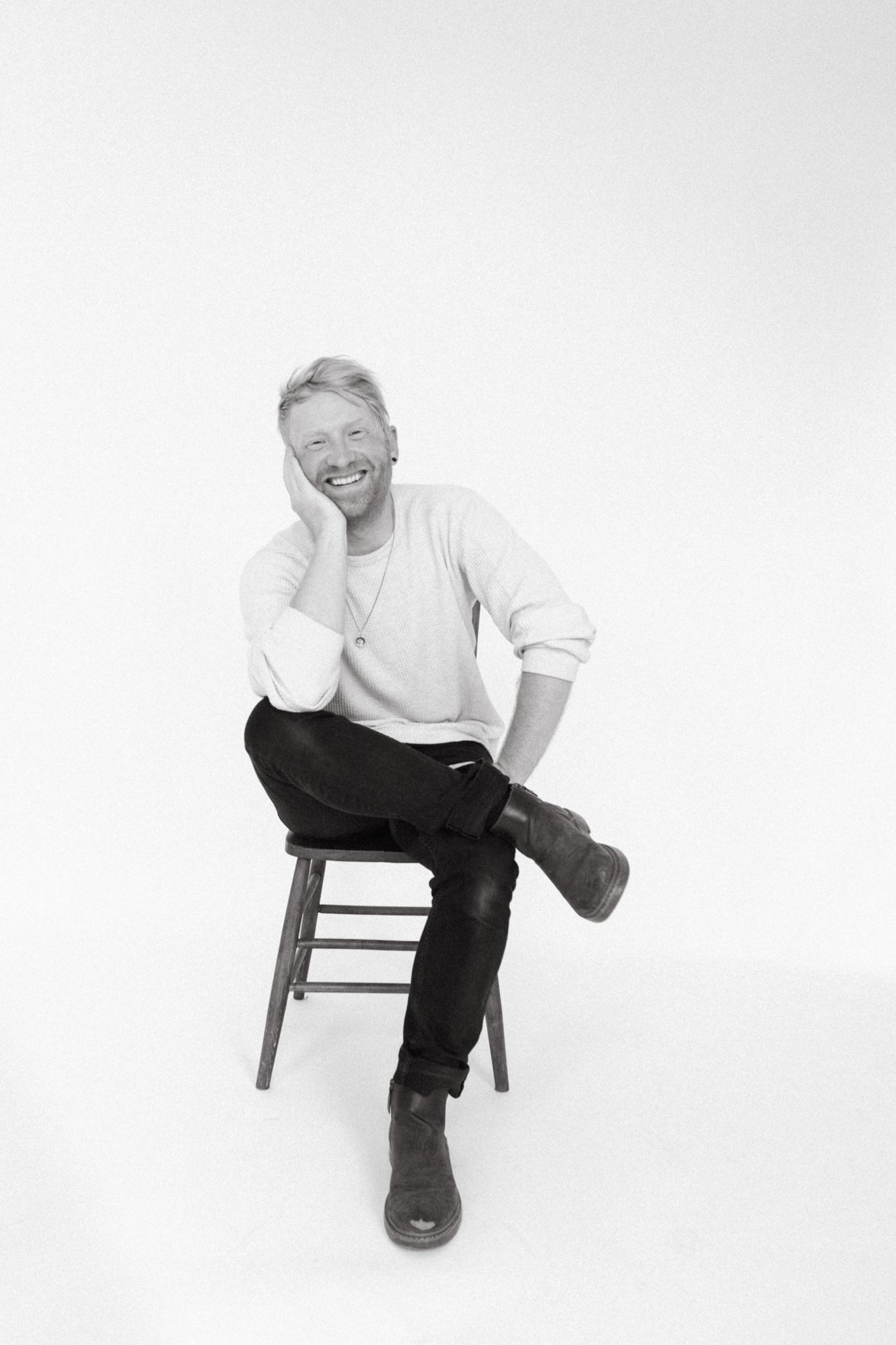 A person in white sits casually on a wooden chair against a white background.