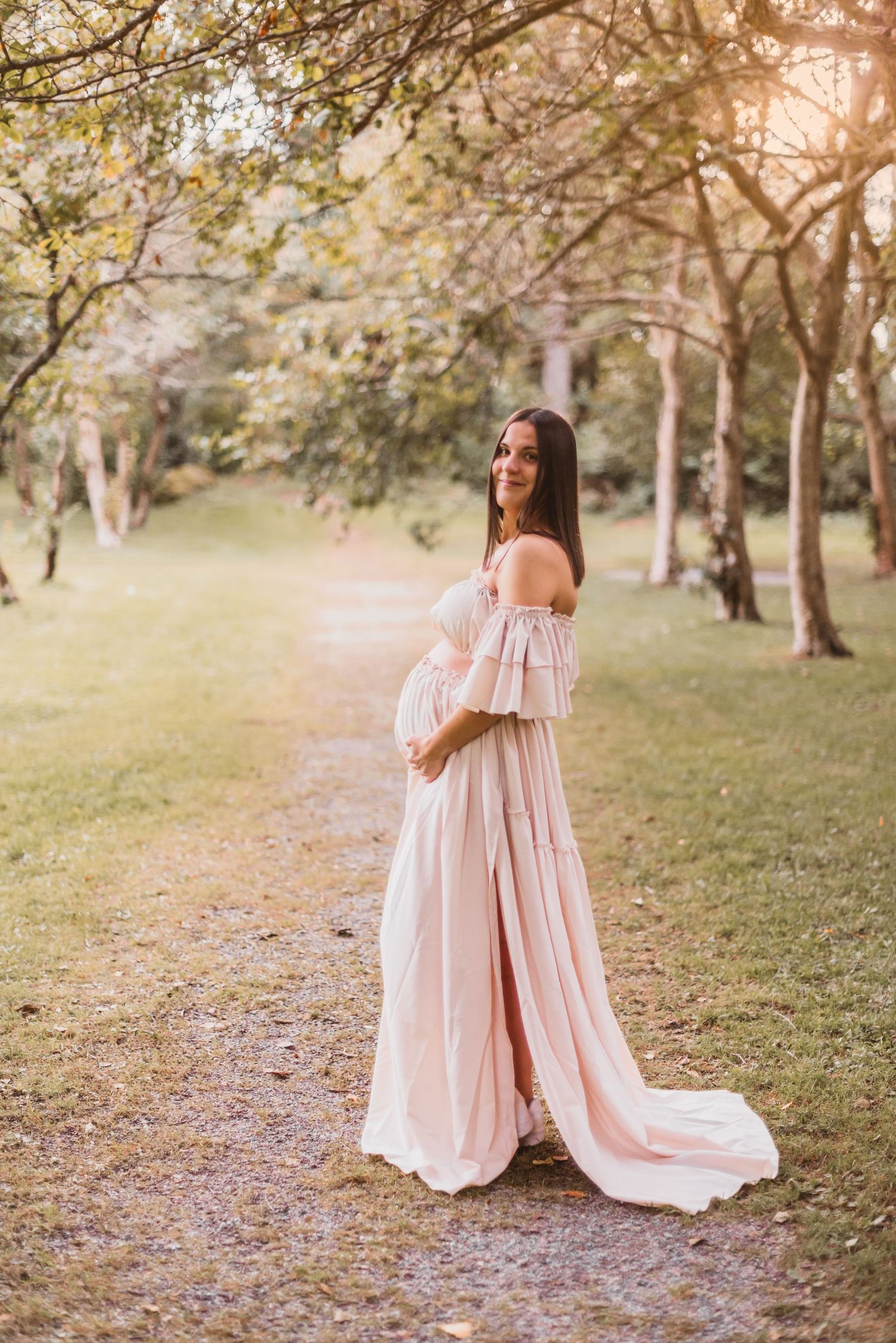 A maternity photo in a flowing white lace dress standing in tall grass and woodland setting.
