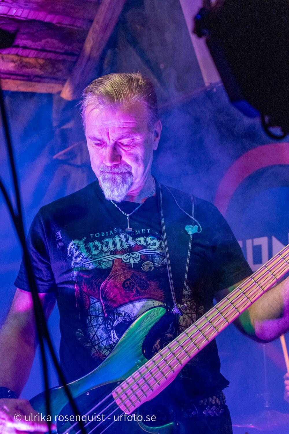 Basistgitarrist uppträder på scen i svart band-t-shirt upplyst av dramatisk lila och blå belysning.