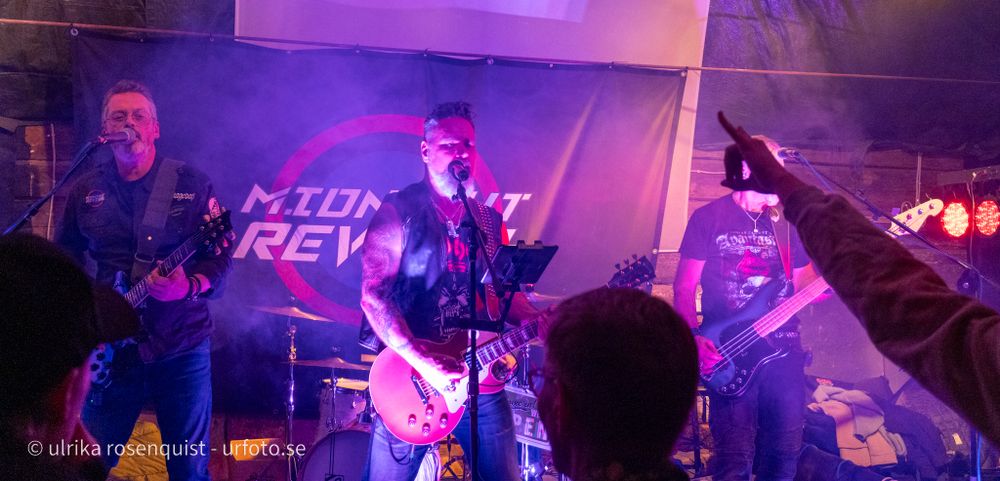 Ett rockband uppträder på scen badande i lila ljus med entusiastiska publiksilhuetter synliga i förgrunden.