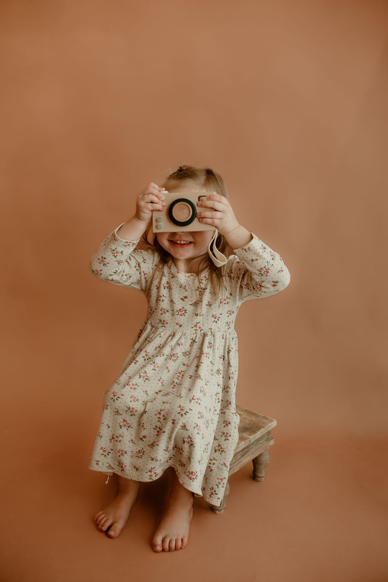 Young child in floral dress holding vintage camera while sitting on wooden stool.