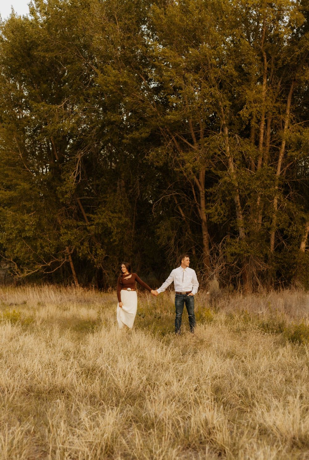 Western Couple walks hand in hand through tall grass field at sunset.