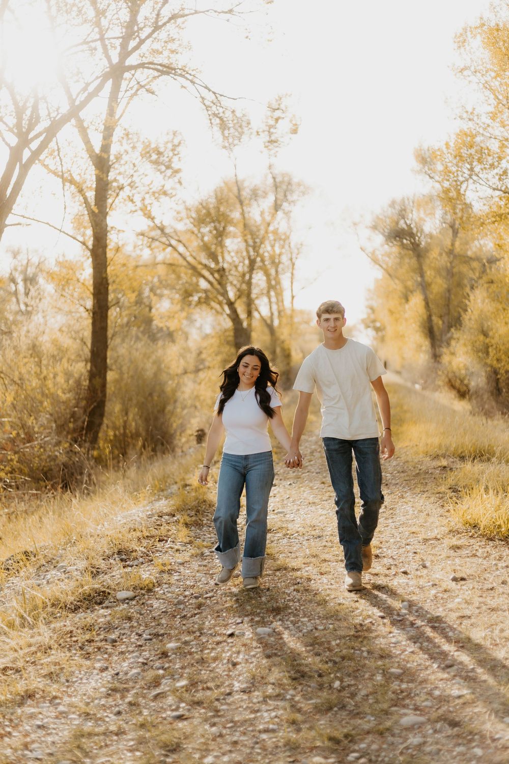 A western couple walks hand in hand along a sunlit dirt path through autumn trees casting long shadows.