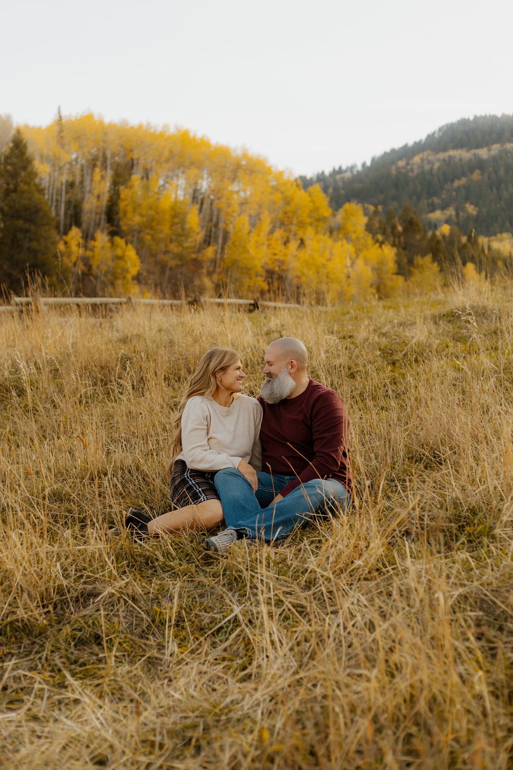 Couple sitting in tall grass during golden hour with fall foliage in background.