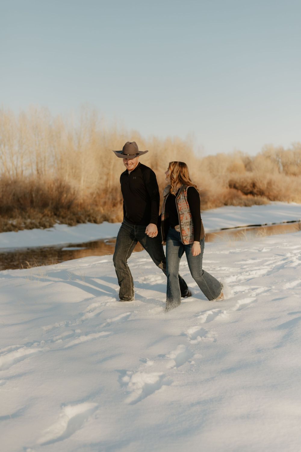 Western Couple walking together on a snowy landscape with winter sunlight and bare trees in the distance. Idaho Beauty