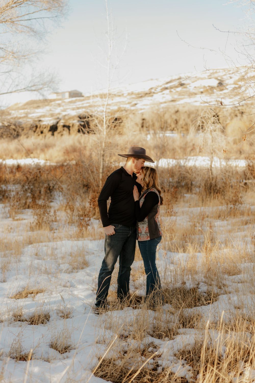 Western Couple standing together in a snowy winter field with dried grass and bare trees.
