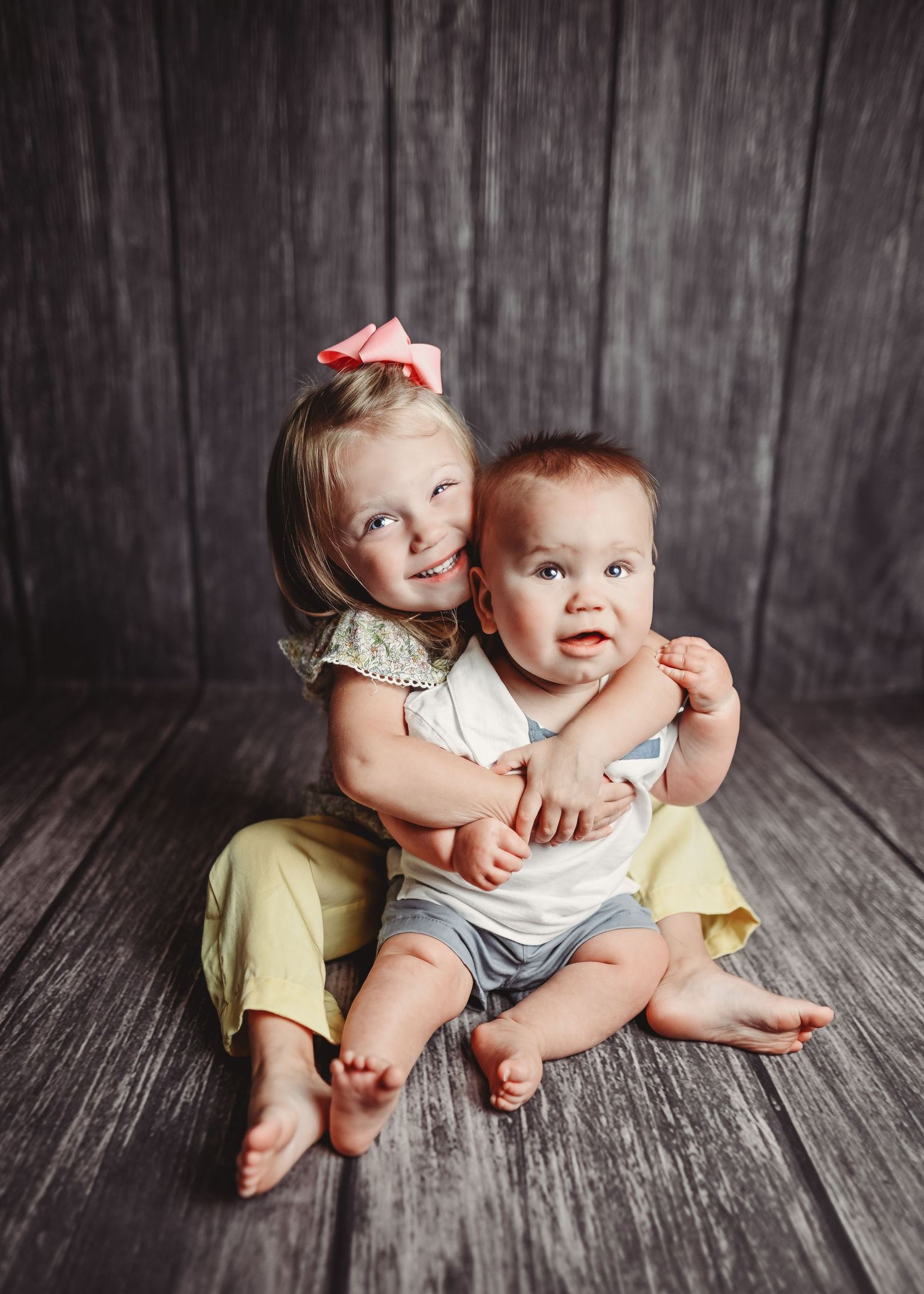 Two small children sitting together on wooden floor with rustic backdrop.