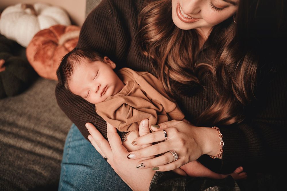 An infant in a brown onesie rests peacefully while being cradled in loving arms at home.