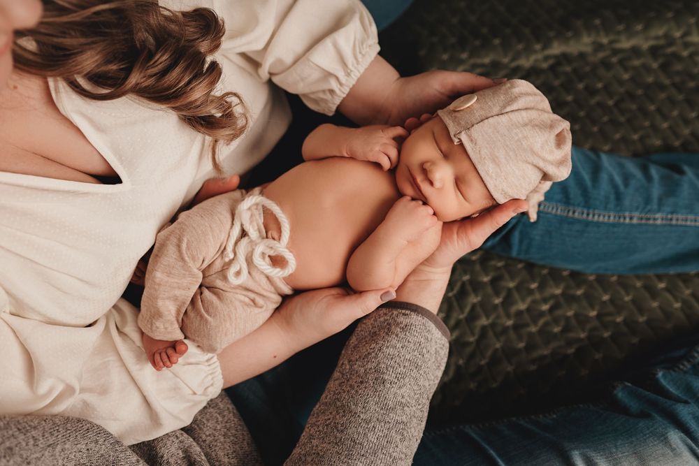 Newborn baby wearing soft beige clothing rests peacefully in their caregiver's arms on a textured green blanket.