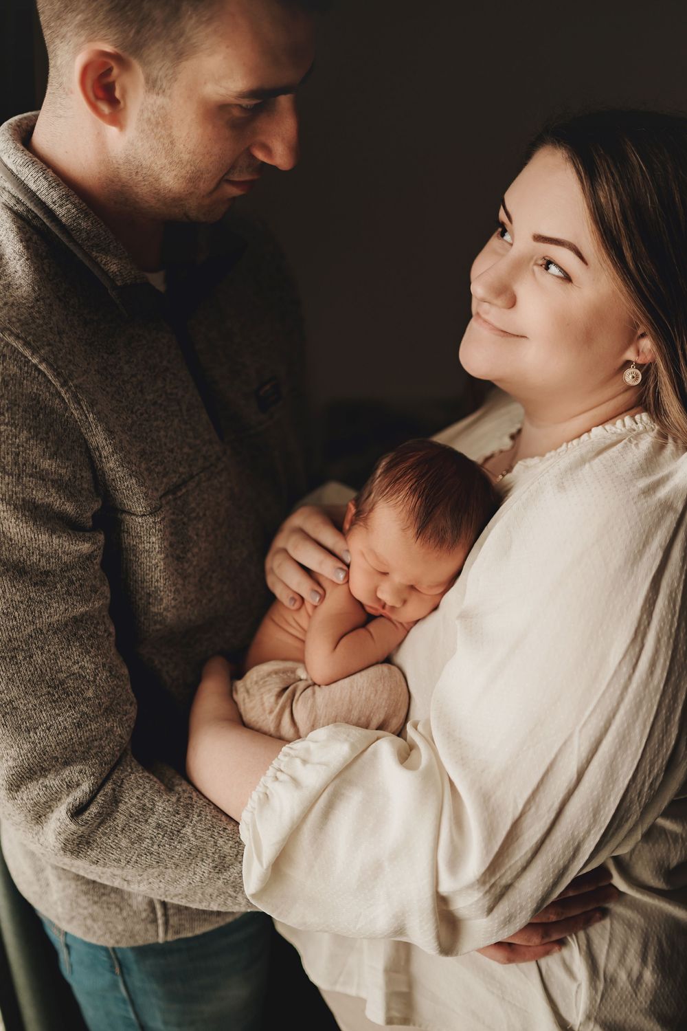 Intimate family moment with sleeping newborn cradled between parents in warm neutral tones against dark background.