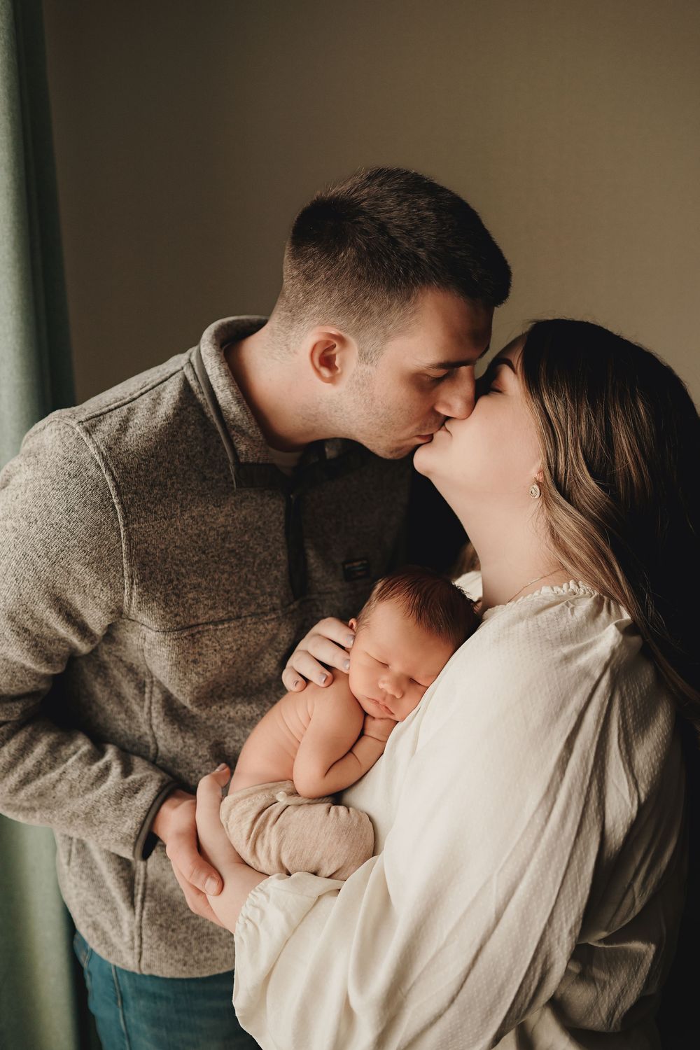 Tender moment between couple sharing affection while holding their sleeping newborn baby in warm lighting.