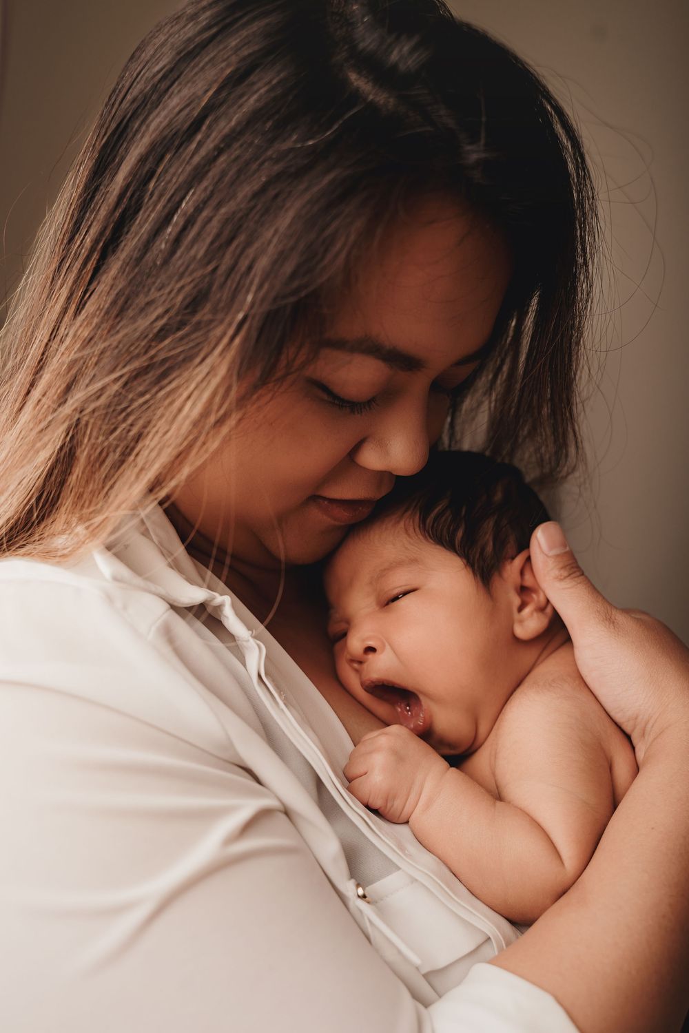 A tender moment between a parent and sleepy newborn baby in warm lighting against a dark background.