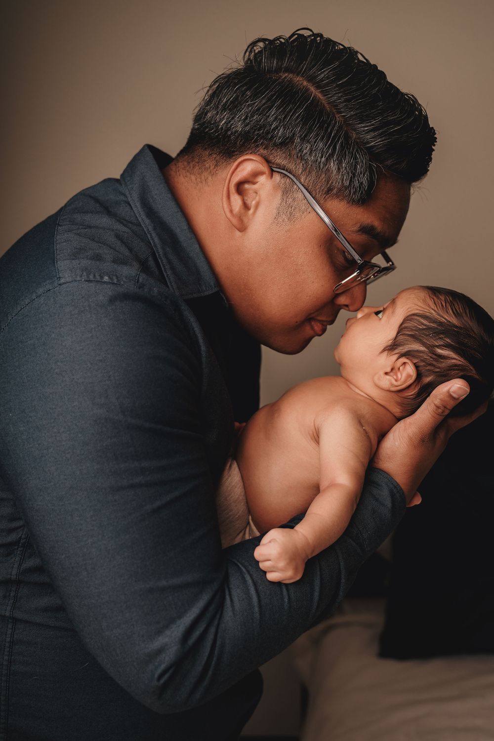 A tender moment captured between parent in dark attire and a shirtless newborn baby against a neutral background.