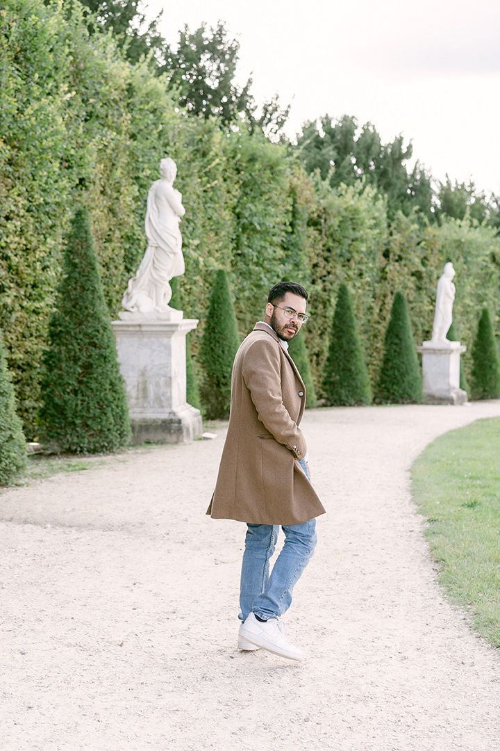 Man in brown coat posing casually on garden path with classical statues and hedges.