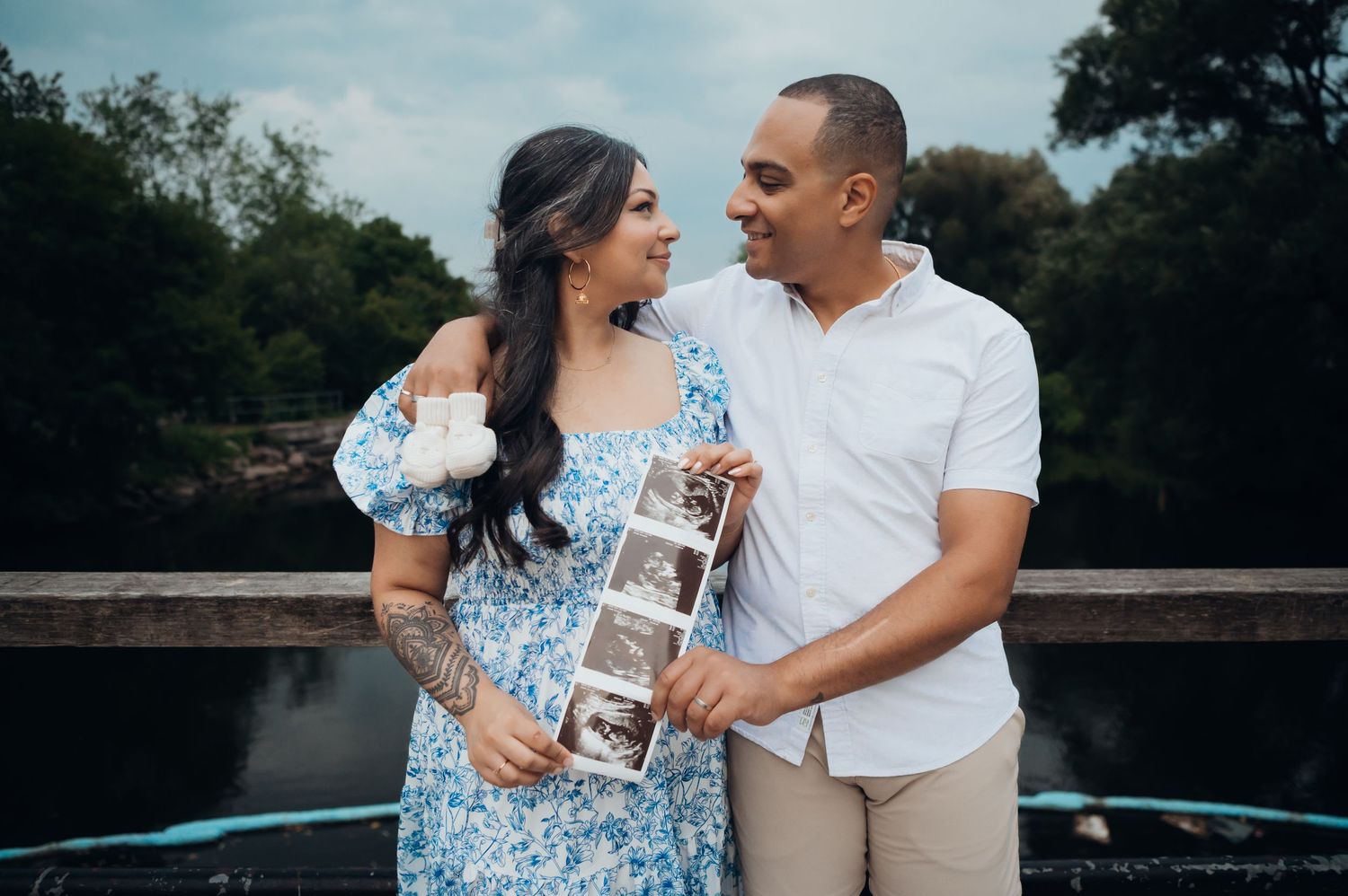 Pregnant couple smiling at each other while holding ultrasound photos outdoors near a scenic lake.