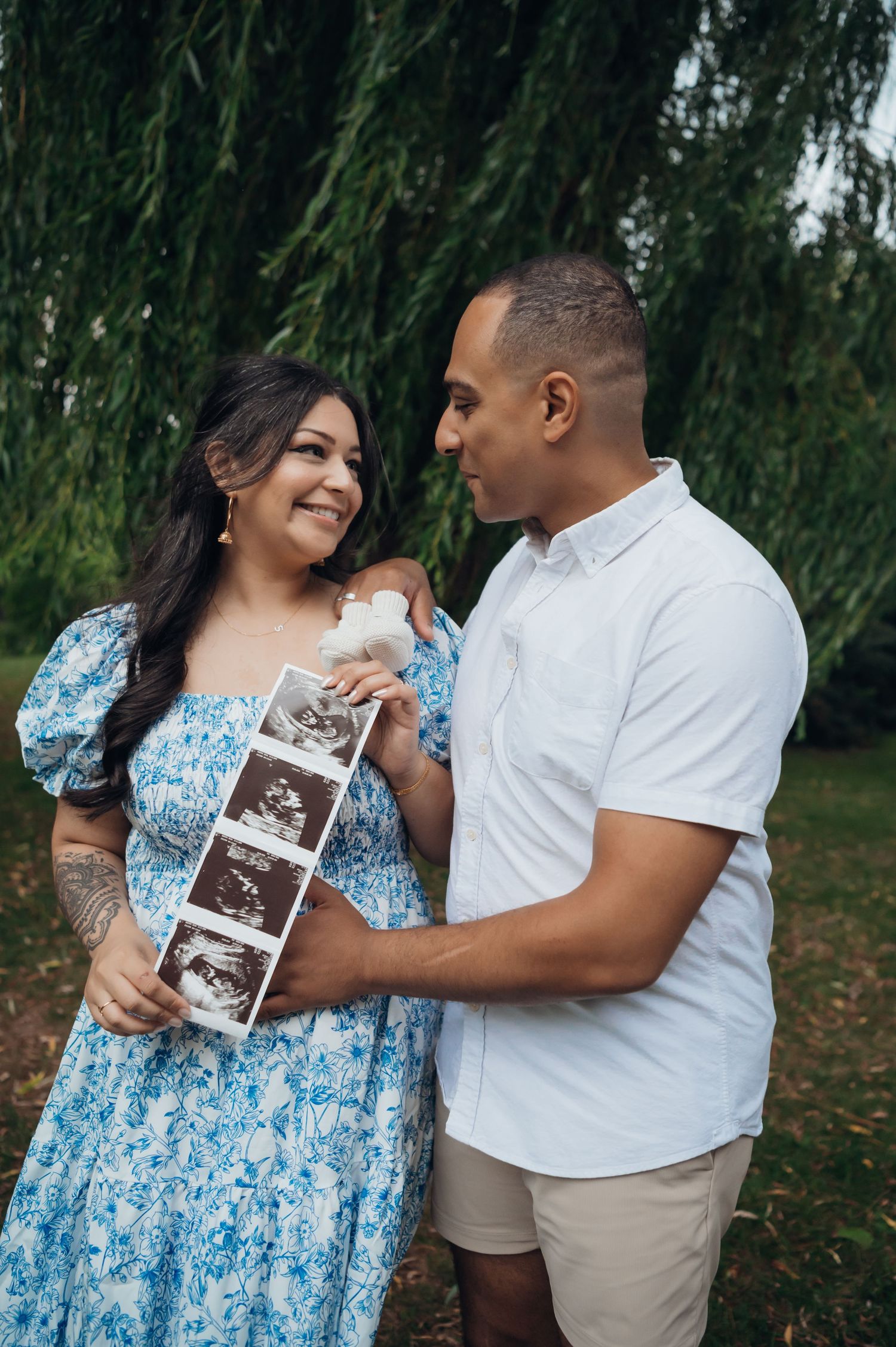 Happy couple holding ultrasound photos outdoors under a willow tree, celebrating their pregnancy together.