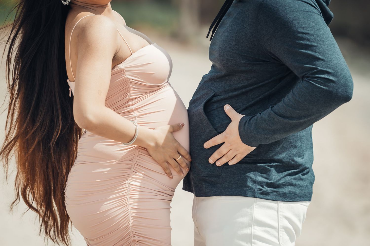 Pregnant woman in pink dress and partner touching baby bump outdoors during maternity photo shoot.