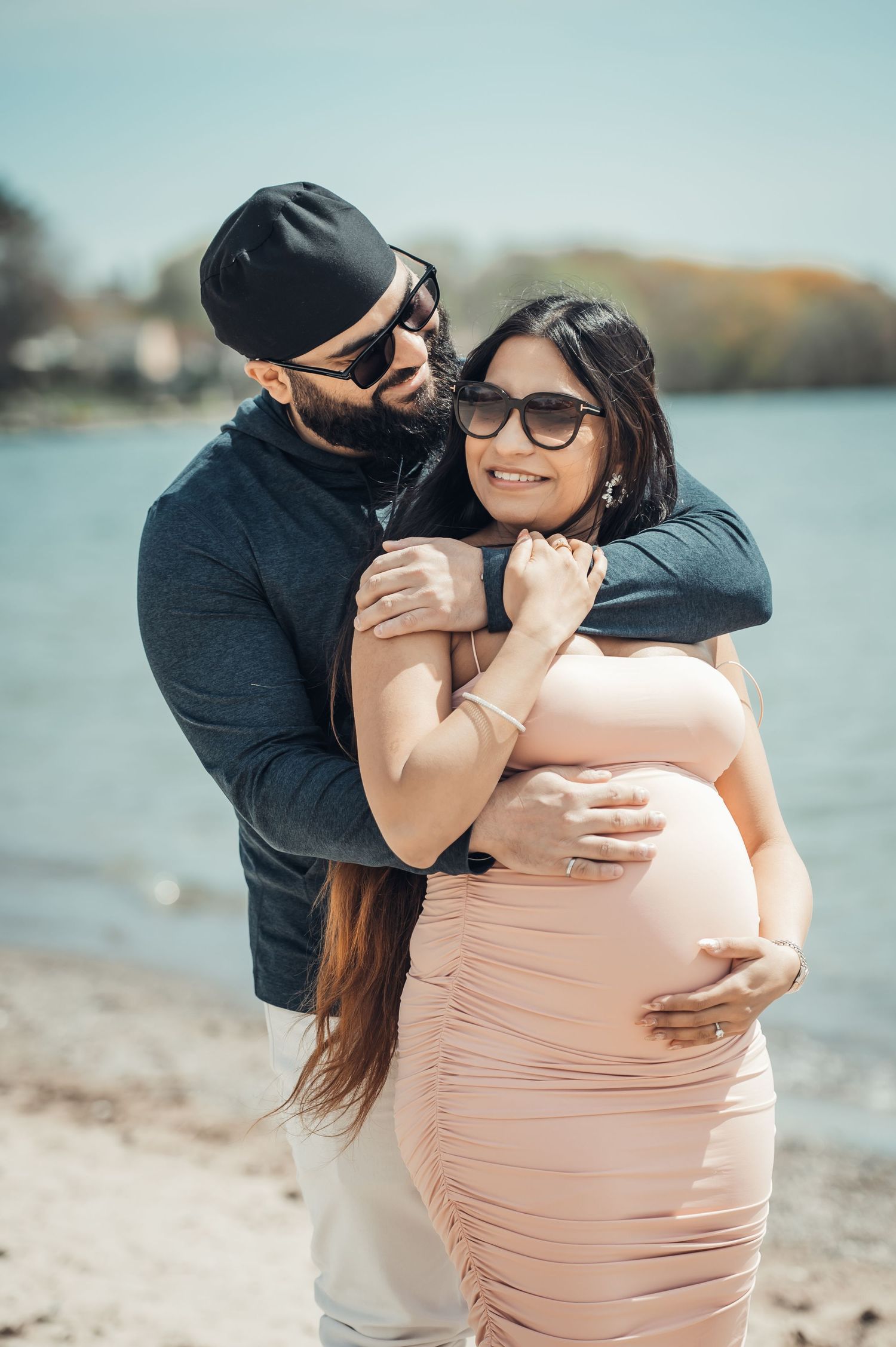 Couple embracing on beach, pregnant woman in pink dress with man in dark outfit, maternity photo shoot by the water.