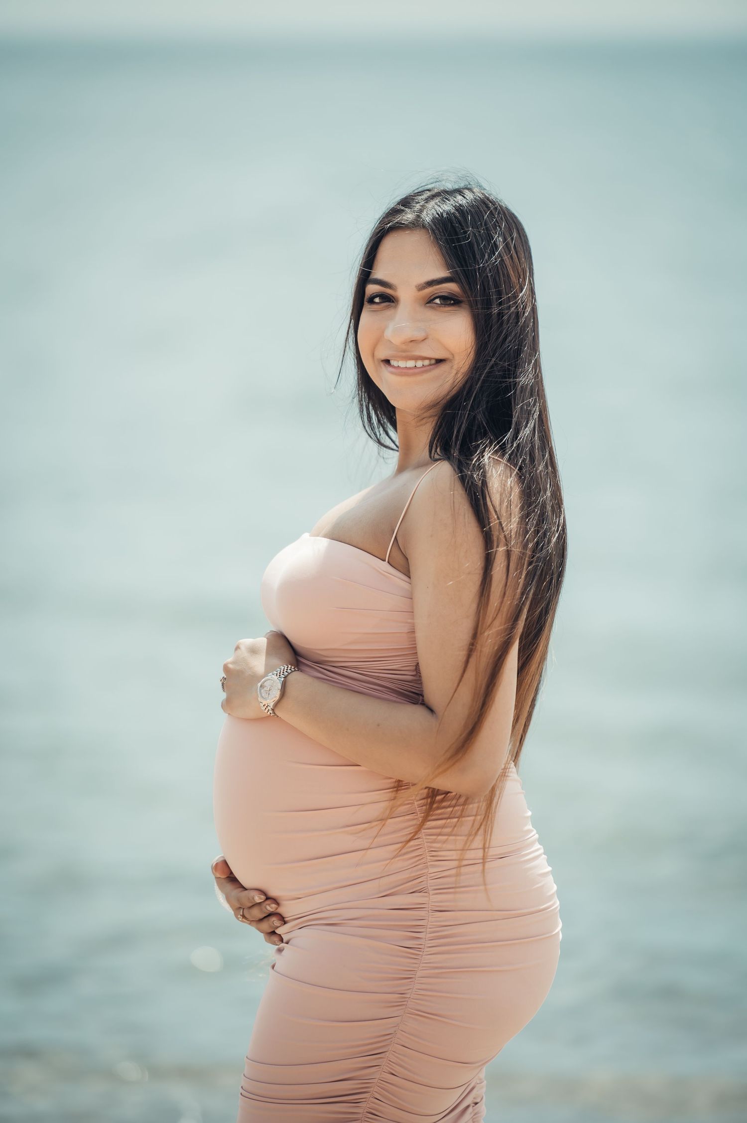 Smiling pregnant woman in pink dress cradling her baby bump on a serene beach.