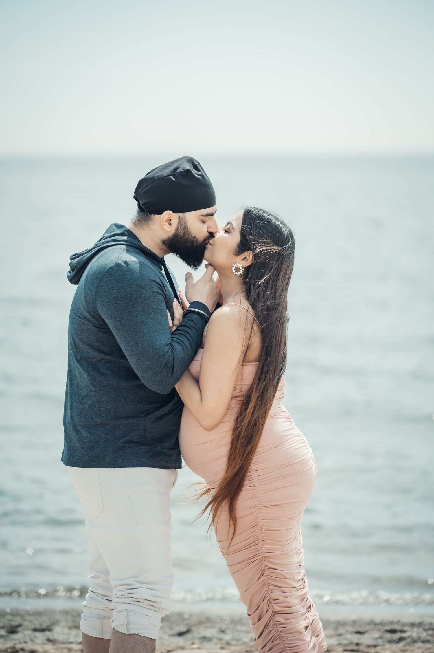 Couple sharing a romantic kiss on a beach, woman in pink dress and man in blue hoodie with dark turban.