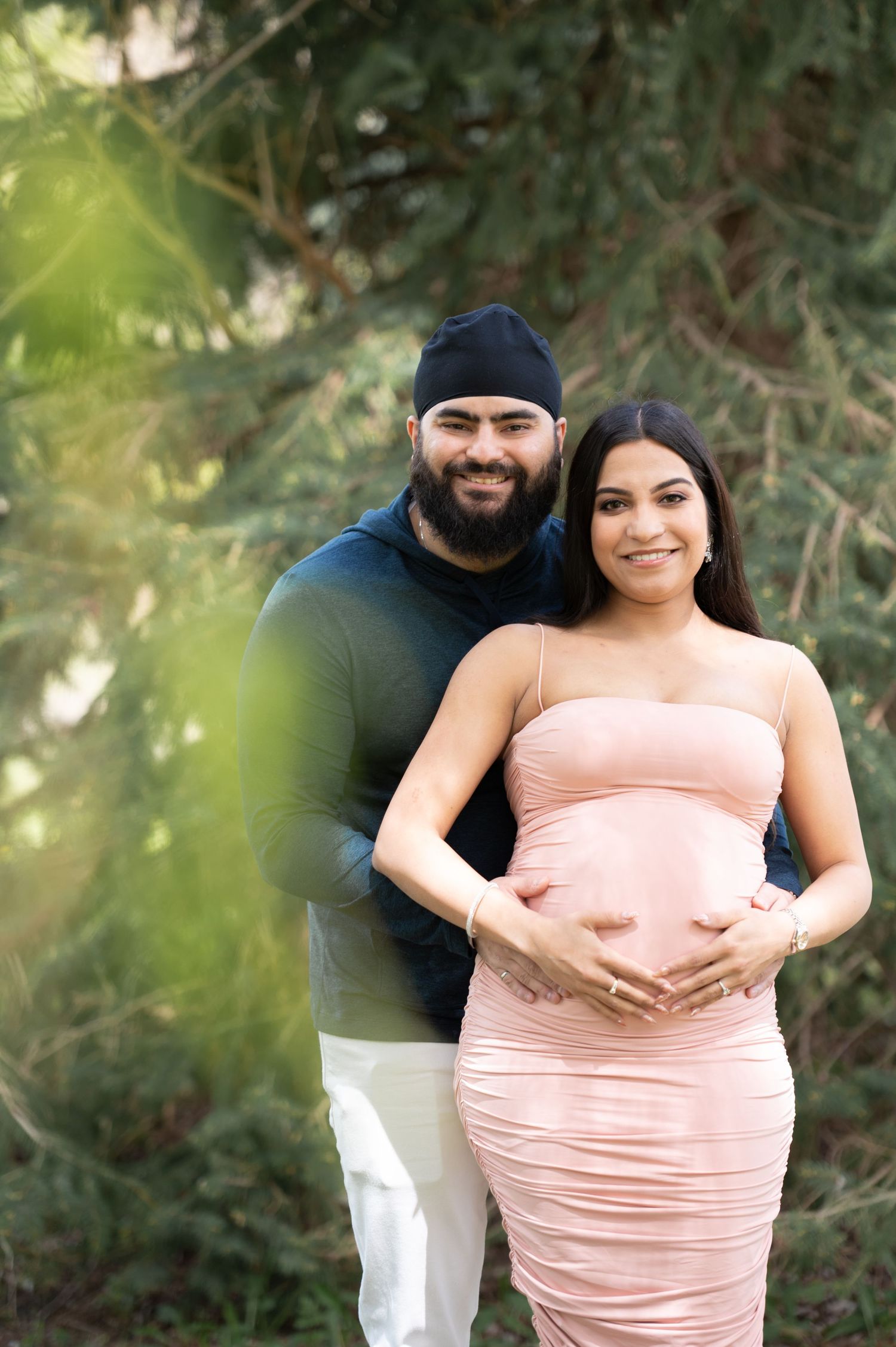 Smiling couple poses outdoors for maternity photo, woman in pink dress cradling baby bump, man in dark outfit standing behind her.