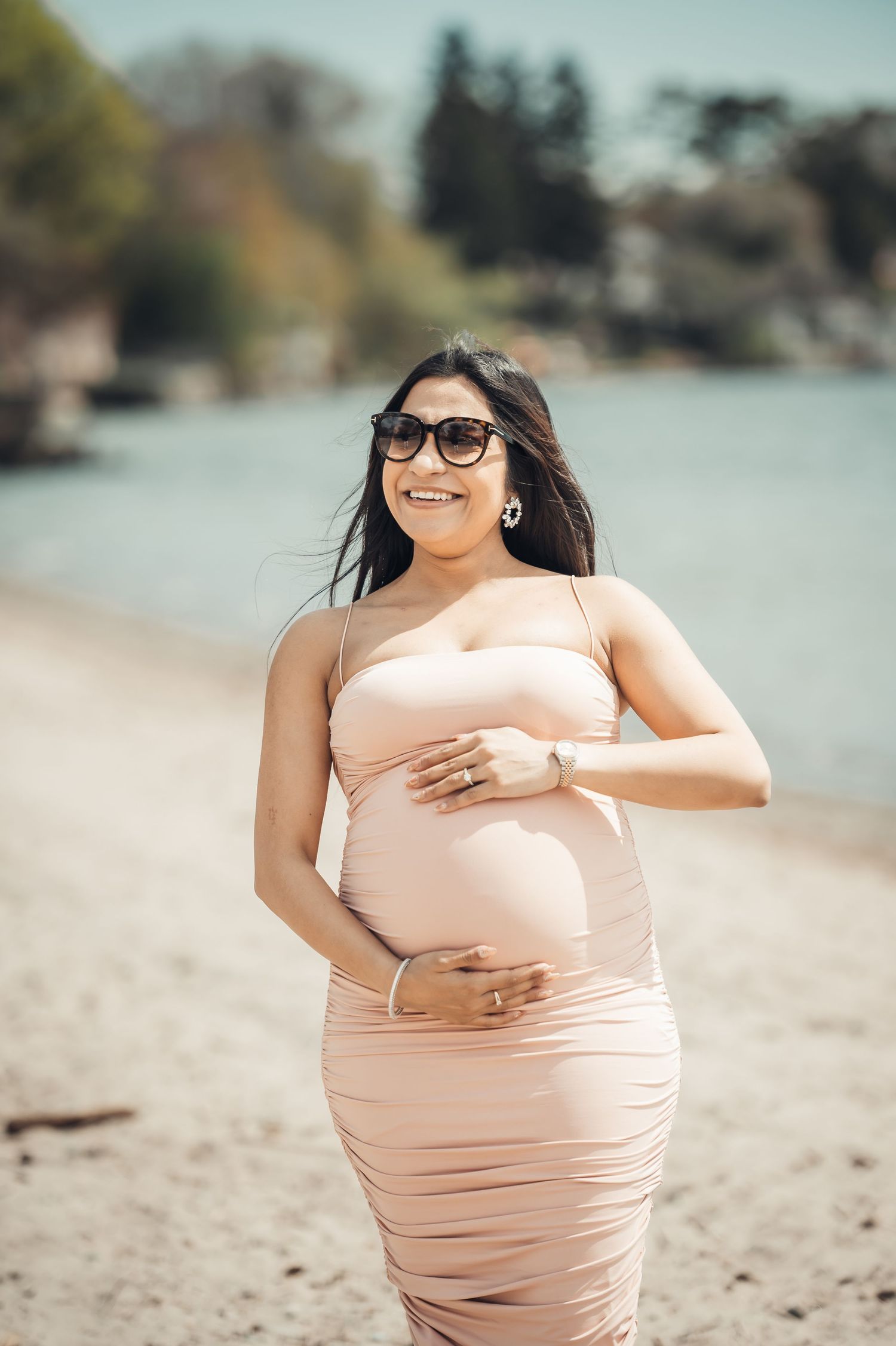 Smiling pregnant woman in pink dress and sunglasses cradling baby bump on sunny beach.
