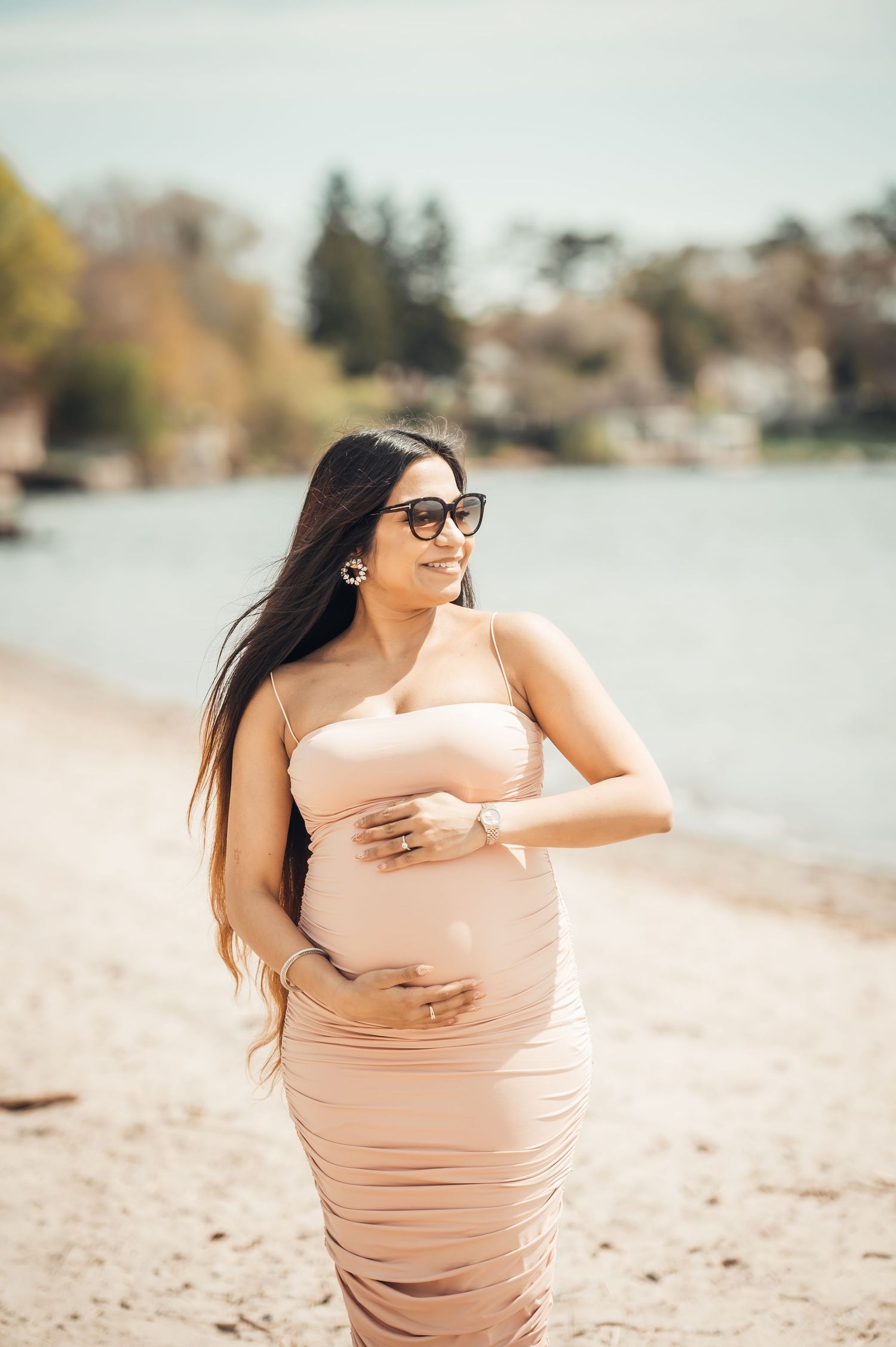 Pregnant woman in blush ruched dress and sunglasses cradling baby bump on sandy beach near lake.
