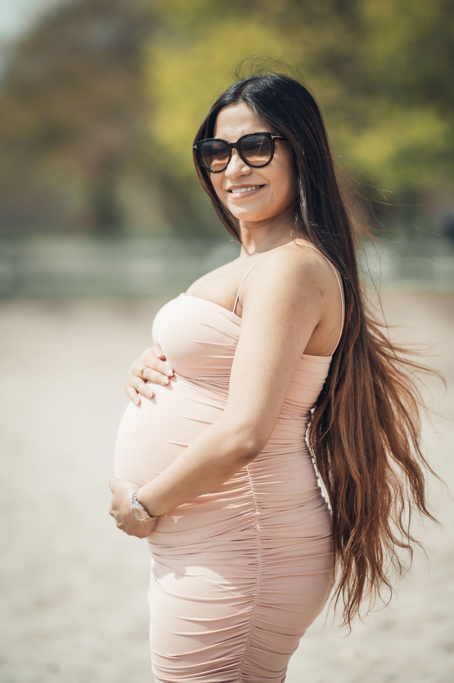 Smiling pregnant woman in pink ruched dress and sunglasses cradling baby bump outdoors on sunny day.