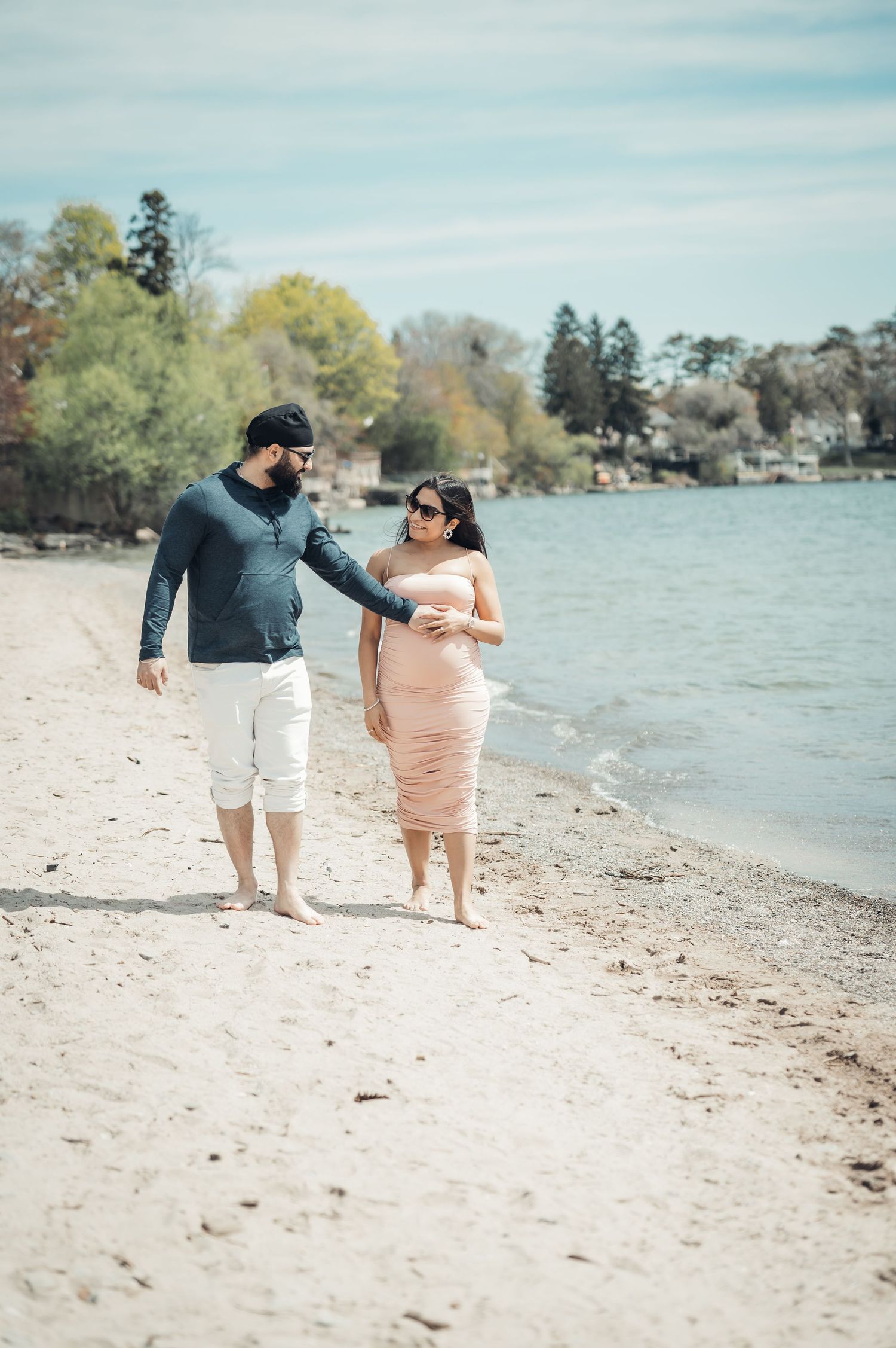 Pregnant couple walking barefoot on sandy beach shore, man in navy shirt and white shorts, woman in pink dress.