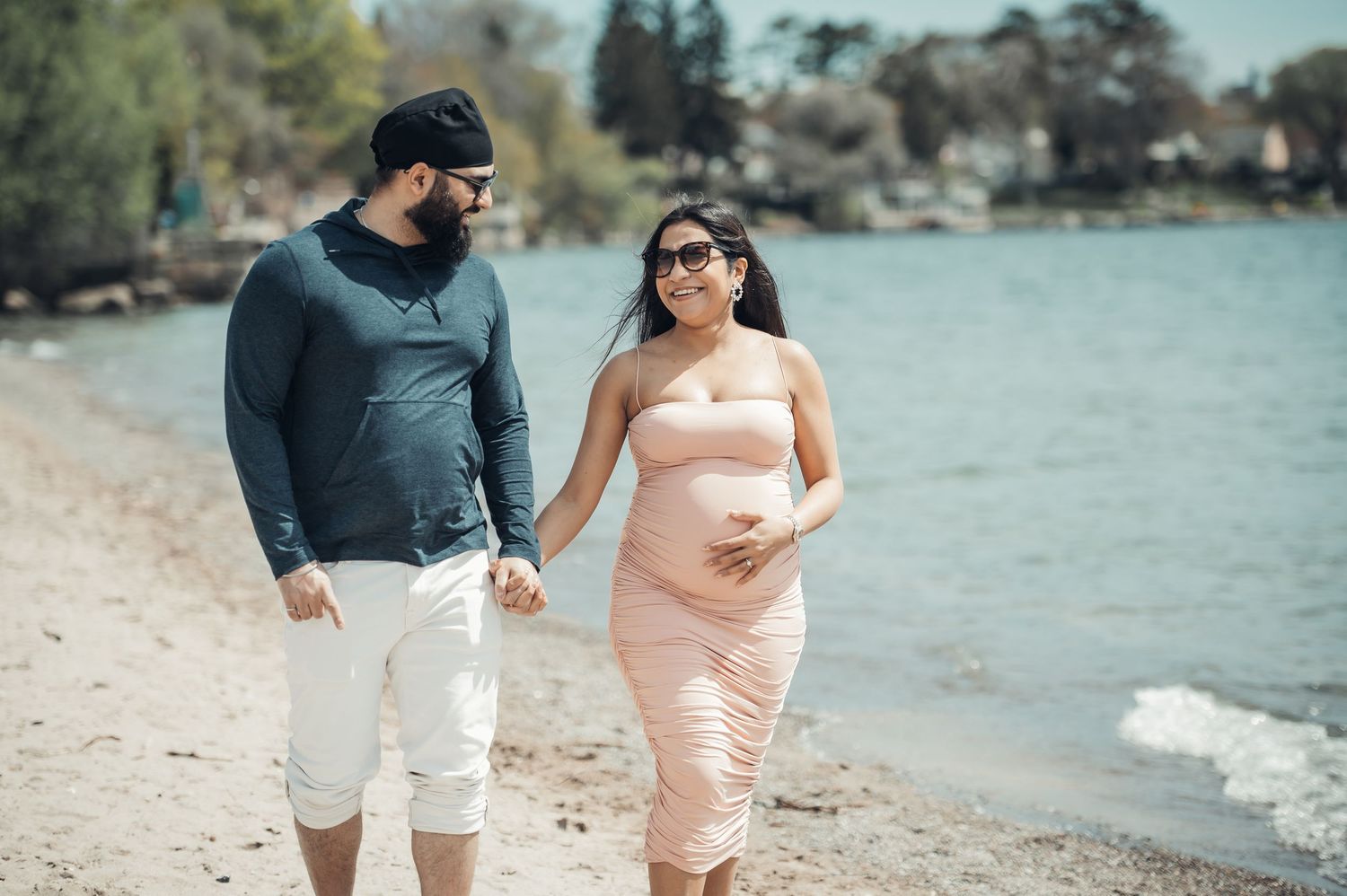 Pregnant couple walking hand in hand on a scenic beach, woman in pink dress cradling baby bump, man in gray shirt.