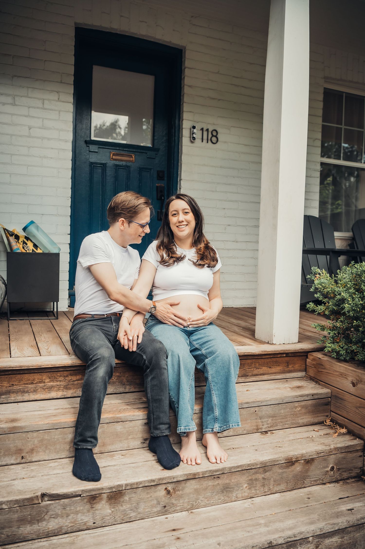 Pregnant couple sitting on wooden porch steps of white brick home, smiling at each other, woman cradling baby bump.