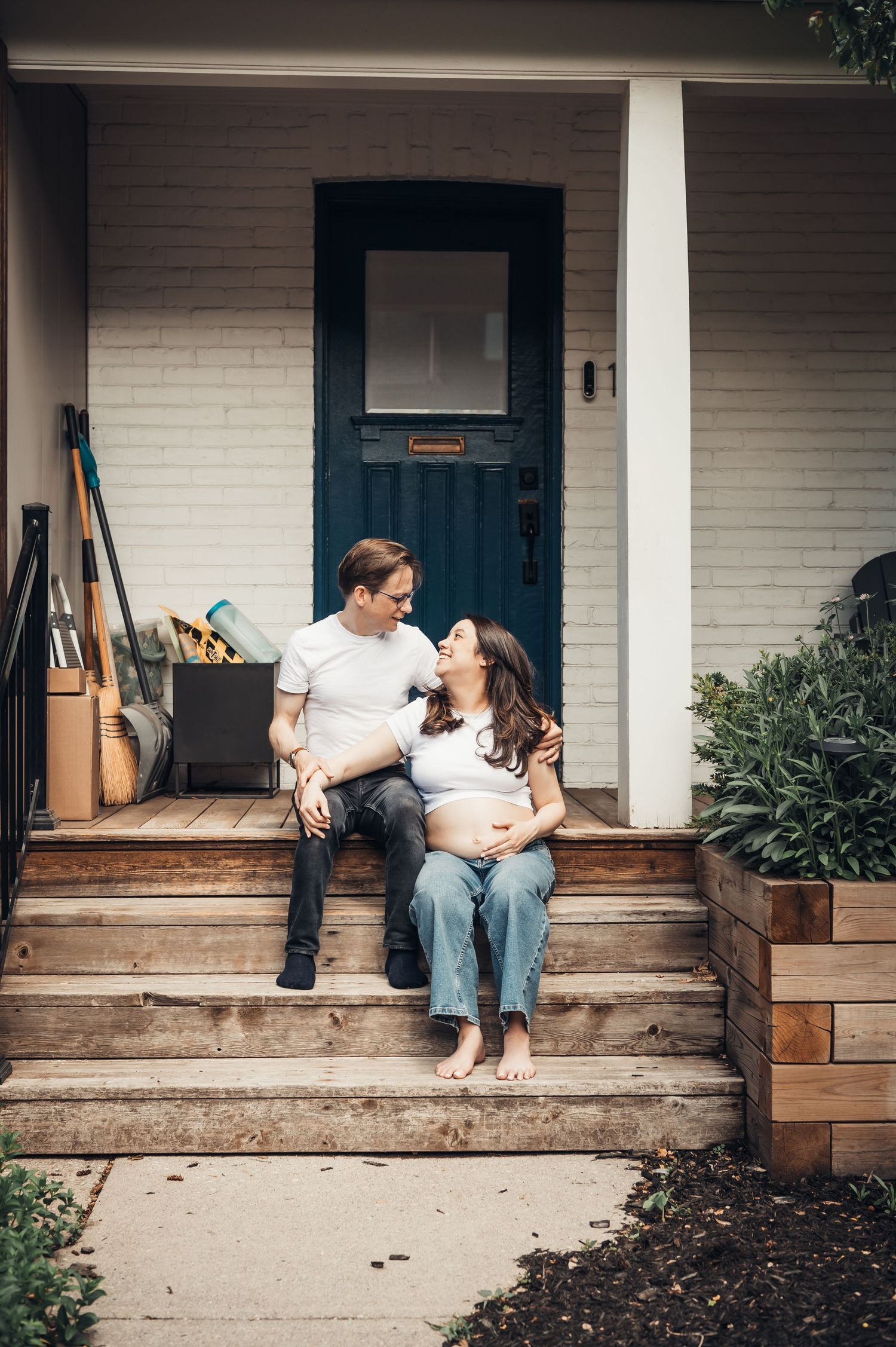 Pregnant couple sitting on front porch steps, embracing and smiling at each other, in casual white tops and jeans.