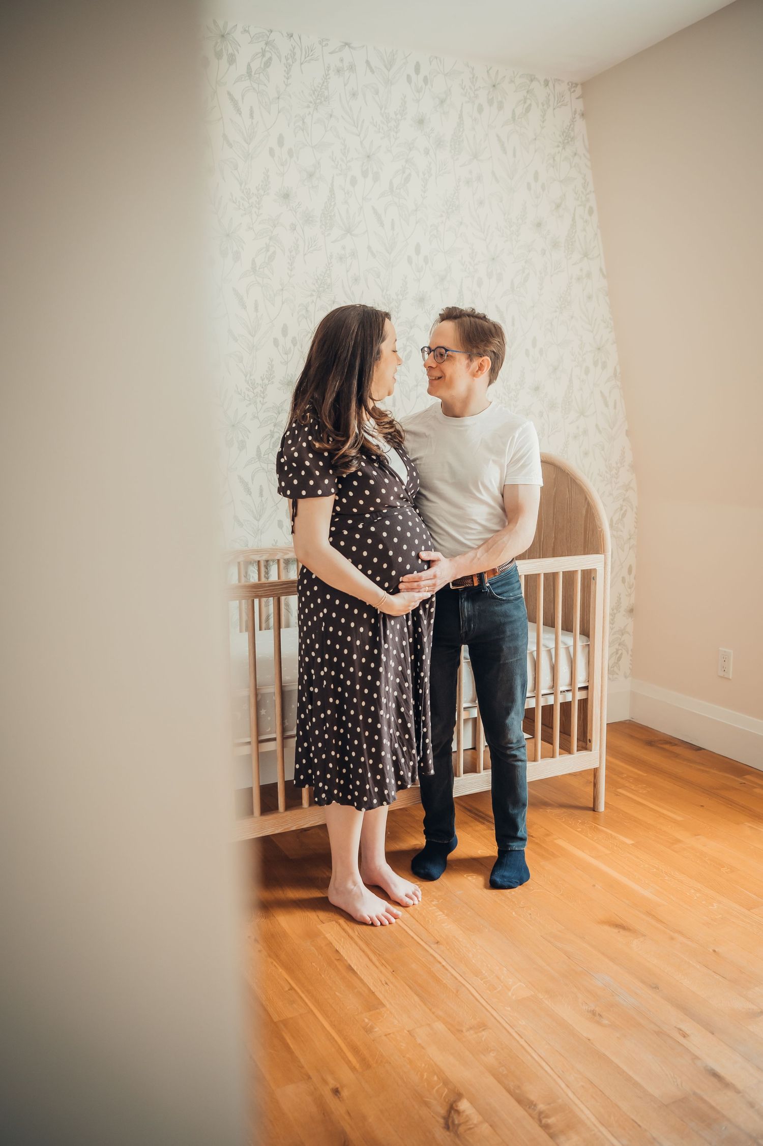 Pregnant couple standing together in a nursery, man holding woman's baby bump, white crib and floral wallpaper in background.