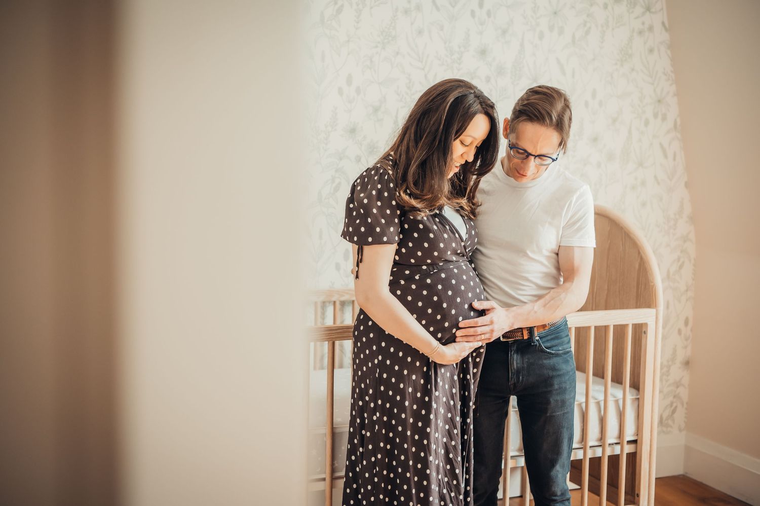 Expectant couple standing by a white crib, looking at baby shoes in a softly lit nursery room.