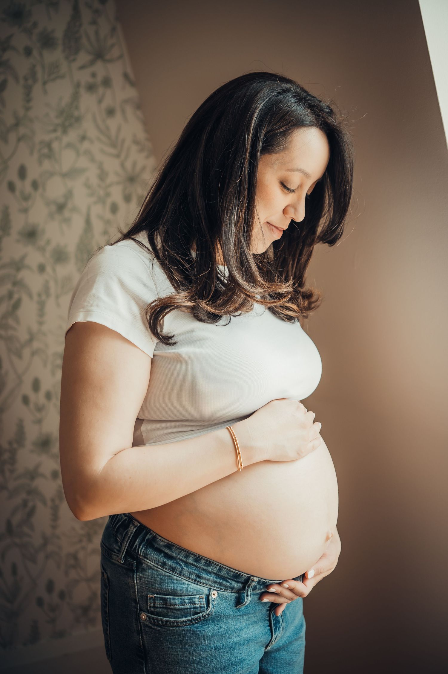 Pregnant woman in white crop top and jeans cradling her baby bump, looking down with a serene expression.