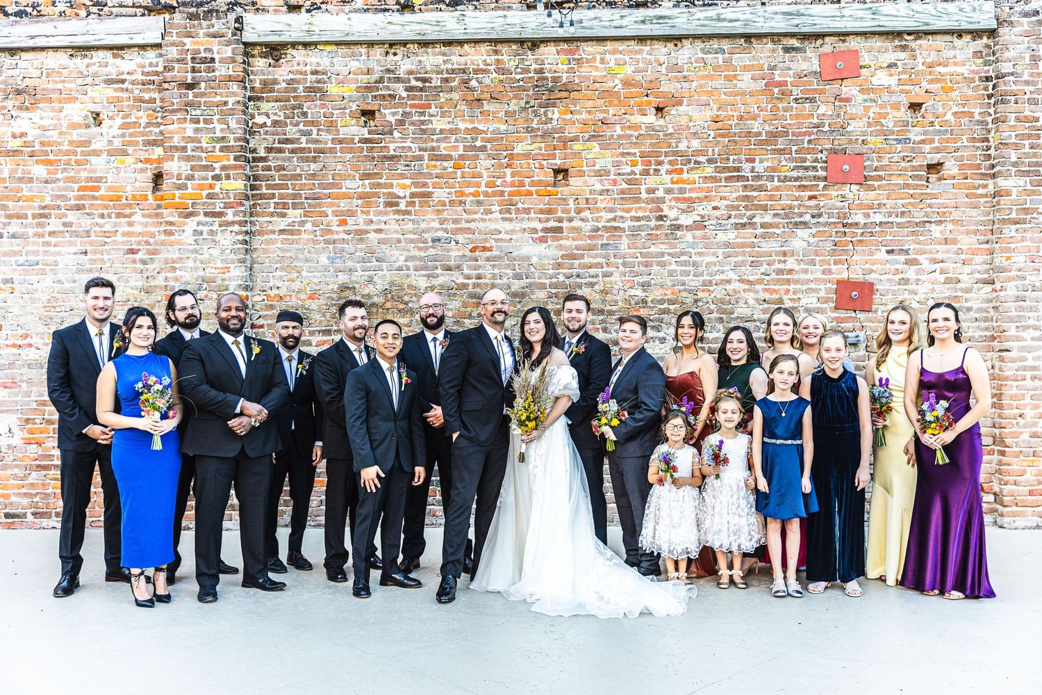 Large wedding party lineup against exposed brick wall with formal attire.