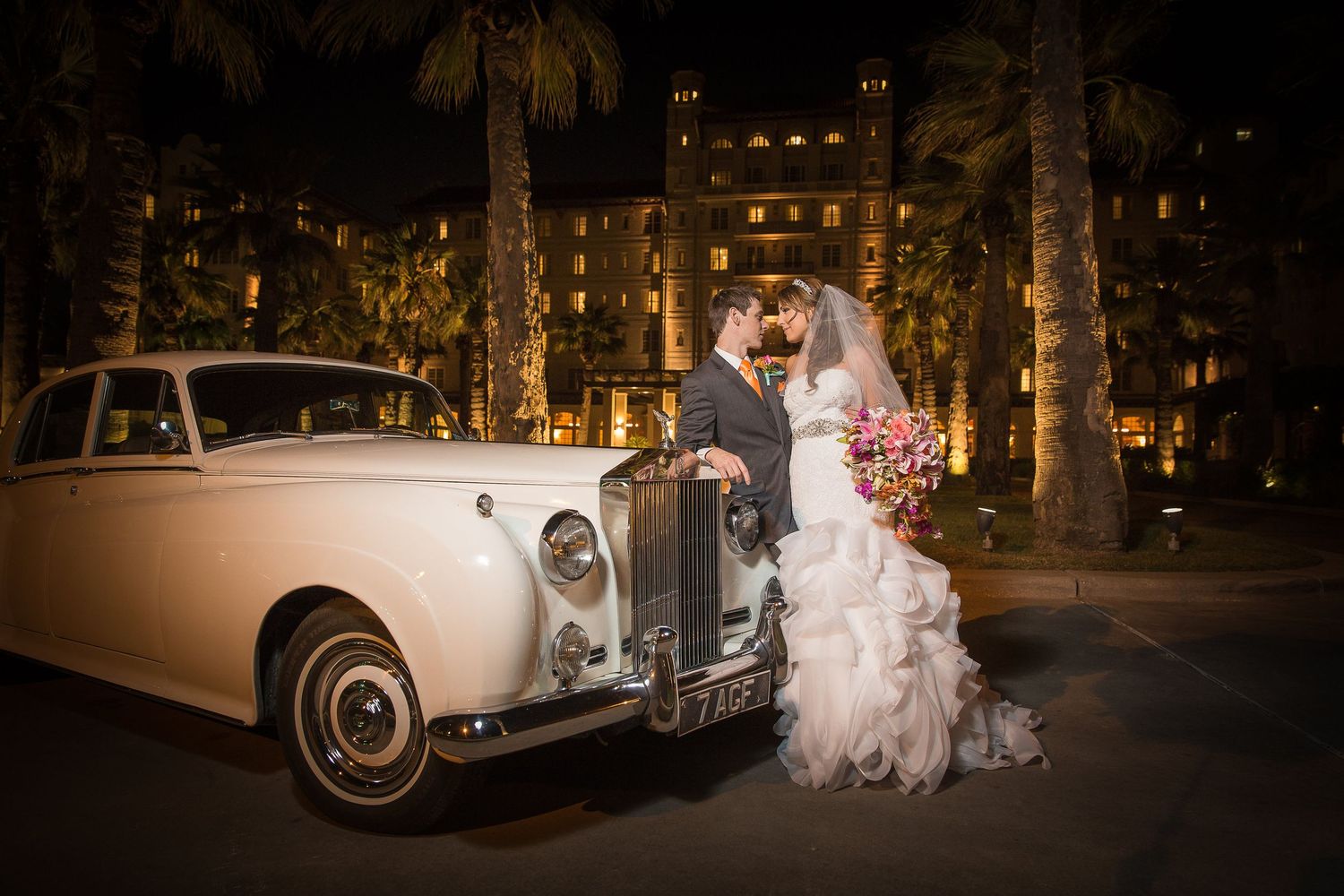 A bride and groom pose at night next to a vintage white Rolls Royce car illuminated by warm lighting.