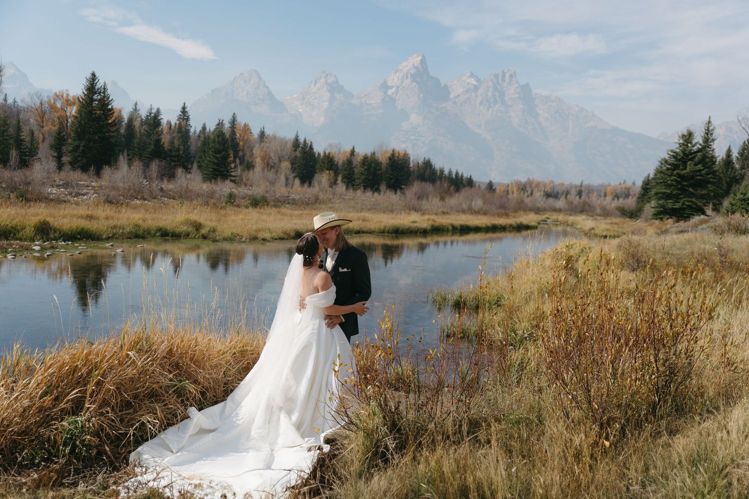 A scenic mountain wedding photo with towering peaks reflected in a tranquil lake surrounded by autumn grass.