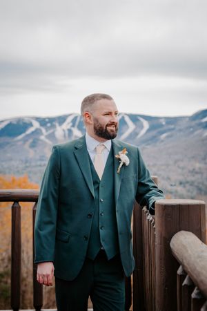 Dapper groom in a dark green three-piece suit with boutonniere posing on a deck with ski mountain backdrop.
