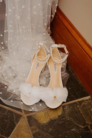 Elegant white bridal heels with pearl ankle straps and bow detail rest on marble floor beside lace veil.