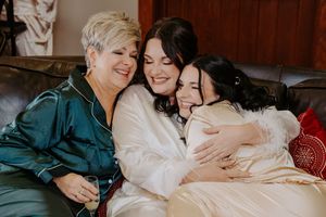 Bride in white robe sharing a joyful hug with mother and bridesmaid on a couch before wedding.