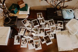 Polaroid photos scattered on rustic wooden table with Instax camera, books, and wedding stationery.