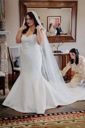 Bride in a white mermaid wedding gown with veil being adjusted by bridesmaid in elegant room.