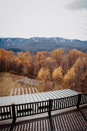 Wooden deck overlooks autumn forest with colorful foliage and snow-capped mountain ski slopes in the background.