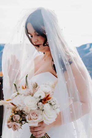 Elegant bride in strapless gown holding white and blush rose bouquet beneath pearl-adorned wedding veil.