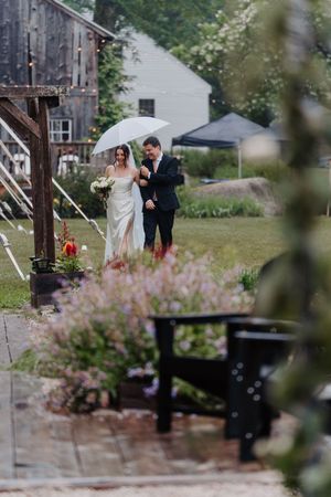Bride and groom walk together under a white umbrella in the rain at a rustic barn wedding venue.